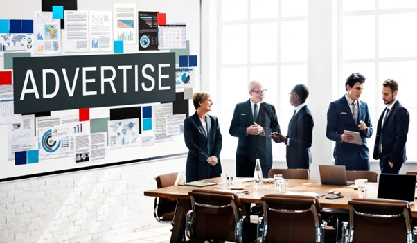  Marketing team discussing advertising strategy in a meeting room with campaign charts and “Advertise” board. 