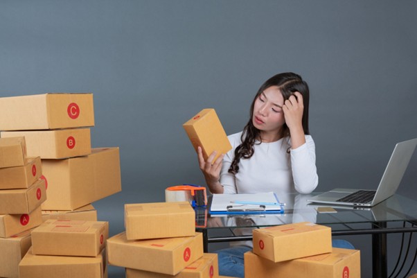 : Woman at a desk surrounded by shipping boxes, looking confused while checking a package, with a laptop and packing supplies nearby.
